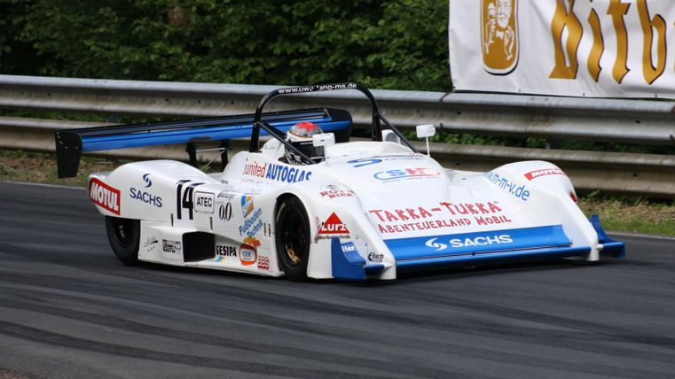 A race car with a striking white and blue design is driving on a winding track. In the background, the safety barriers and a banner are visible.