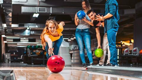 A group of friends is bowling at a modern bowling alley. The woman in the foreground is throwing a red bowling ball while her friends watch closely.