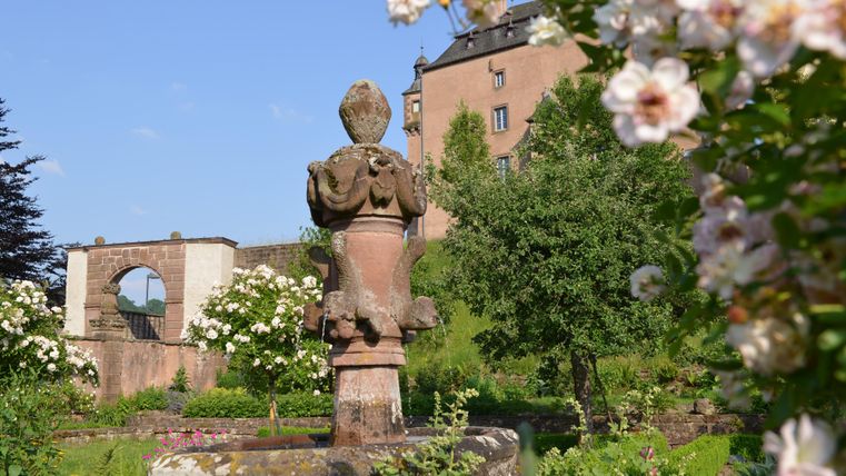 A beautiful garden with a statue in the foreground. In the background, a historical building and blooming roses can be seen.