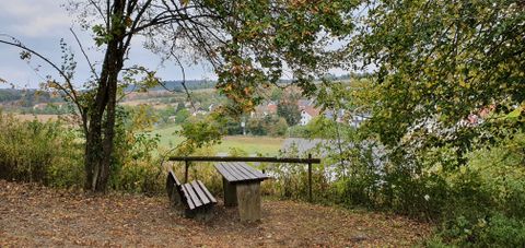Eine Bank unter einem Baum mit Blick auf ein Dorf in einer hügeligen Landschaft.