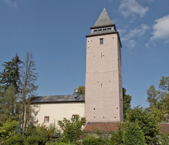 Der Bergfried von Kyllburg erhebt sich vor einem klaren blauen Himmel, umgeben von grünen Bäumen und einem Gebäude mit Schieferdach., © TI Bitburger Land