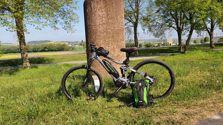 Fahrrad lehnt an einer Steinsäule auf einer Wiese, umgeben von Bäumen und Landschaft.