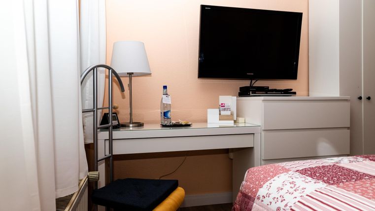 Ein gemütliches Zimmer mit einem Fernseher an der Wand und einem Schreibtisch. Neben dem Schreibtisch steht eine Lampe und eine Flasche Wasser.