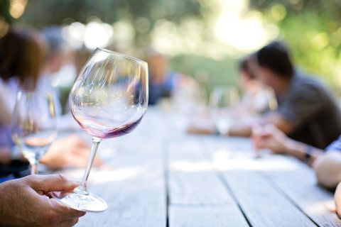 A wine glass with red wine is being held in the hand. Blurry people can be seen in the background at a long table.