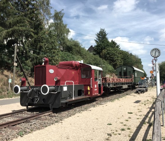 Rote Lokomotive und alte Waggons auf Gleisen am alten Bahnhof in Pronsfeld, umgeben von Bäumen und Blumen., © Eifel Tourismus GmbH