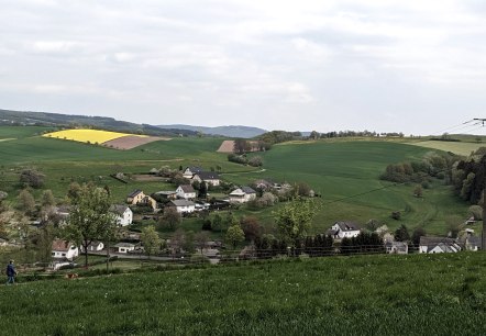 Grüne Hügel mit gelbem Rapsfeld, ein Dorf im Tal und ein Spaziergänger im Vordergrund. Bewölkter Himmel., © A. Girards