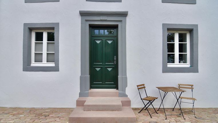 A green front door with a step and two windows beside it. In front of the door is a small table with two chairs.