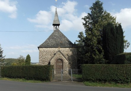 Stone chapel with pointed roof and weathercock, surrounded by trees and hedges, against a blue sky., © Conny Meier