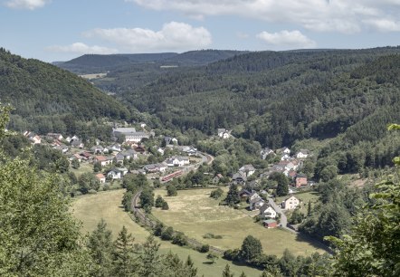 Panoramablick auf ein Dorf in einem grünen Tal, umgeben von bewaldeten Hügeln und Wiesen. Ein Zug fährt durch das Dorf., © Rudolf Höser
