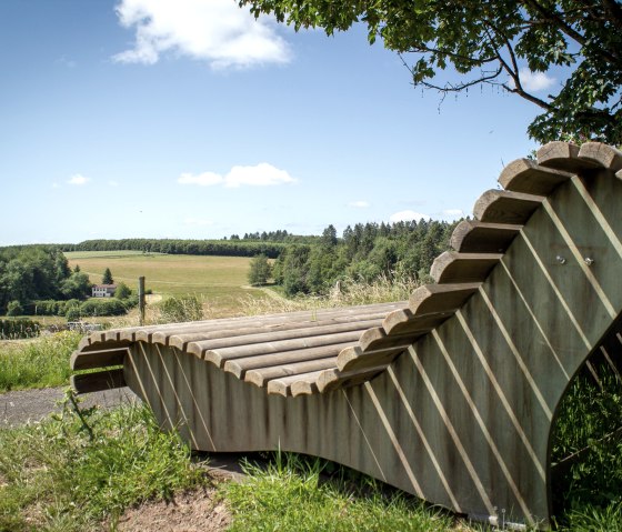 Banc de repos en bois avec vue sur des champs et des forêts verdoyants sous un ciel bleu à Neidenbach., © TI Bitburger Land - Monika Mayer