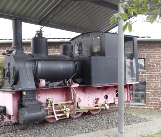 An old steam locomotive stands under a canopy in front of a brick building. The locomotive is painted black and pink., © Eifelverein Ortsgruppe Speicher