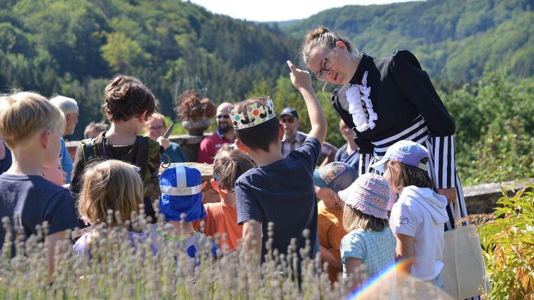 Een groep kinderen luistert aandachtig naar een vrouw in kostuum. Op de achtergrond zijn groene bossen en een zonnig landschap te zien.