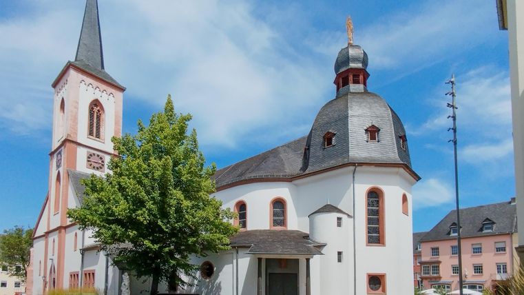 Eine schöne Kirche mit einem hohen Turm und einer markanten Kuppel. Umgeben von Bäumen und einem klaren Himmel.