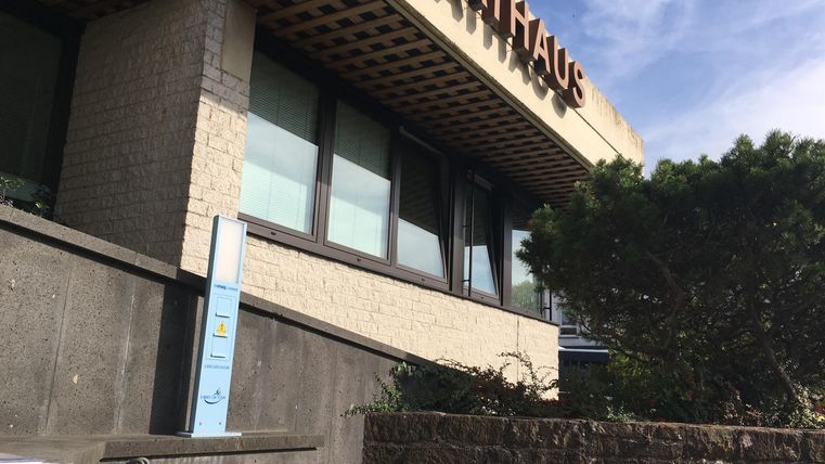 Ein modernes Rathausgebäude mit großen Fenstern und einem Schriftzug. Im Vordergrund sind Sträucher und eine Treppe zu sehen.