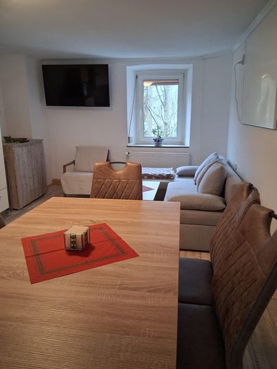 Ein gemütlicher Wohnraum mit einem Holztisch und einem Fernseher an der Wand. Der Raum ist hell und hat große Fenster mit Blick ins Freie.