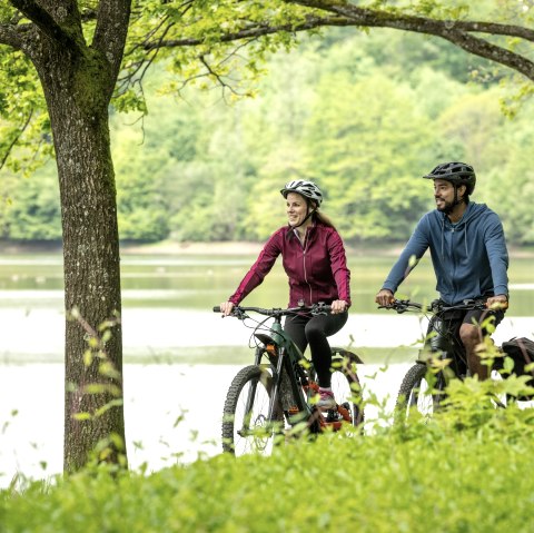 Deux cyclistes roulent en souriant sur un chemin le long du lac de barrage de Bitburg, entourés d'une nature verte et d'arbres., © Eifel Tourismus GmbH, Dominik Ketz