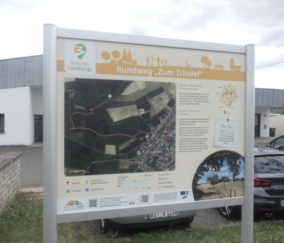 Starttafel des Bitburger LandGang Rundwegs 'Zum Trindel' mit Karte und Infos. Schilder weisen auf den 3,9 km langen Rundweg hin. Autos im Hintergrund., © TI Bitburger Land