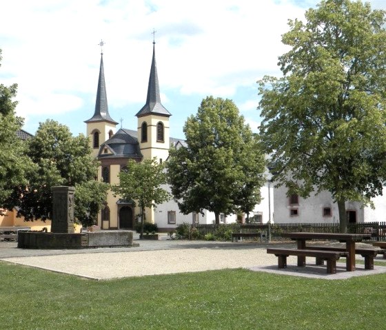 Ein idyllischer Dorfplatz in Idenheim mit einer Kirche im Hintergrund, umgeben von Bäumen. Im Vordergrund sind Bänke und ein Brunnen zu sehen., © Ingrid Penning