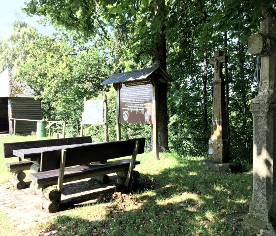 Aire de repos avec des bancs en bois, un panneau d'information et une croix en pierre sous les arbres. En arrière-plan, une cabane en bois et de la végétation verte., © Tourist-Info Bitburger Land M.Mayer