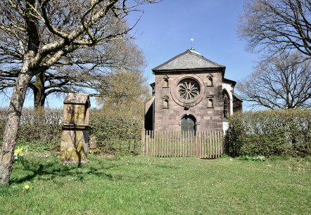 De Frohnertkapel in Oberkail, omringd door bomen en een oude grafsteen op de voorgrond, bij zonnig weer., © TI Bitburger Land