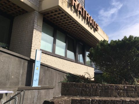 Ein modernes Rathausgebäude mit großen Fenstern und einem Schriftzug. Im Vordergrund sind Sträucher und eine Treppe zu sehen.