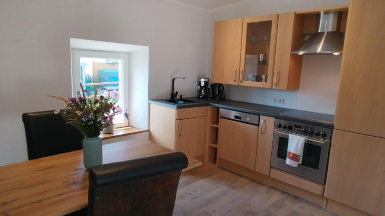 Eine moderne Küche mit Holzschränken und Edelstahlgeräten. Am Tisch stehen frische Blumen und ein Fenster bietet Tageslicht.