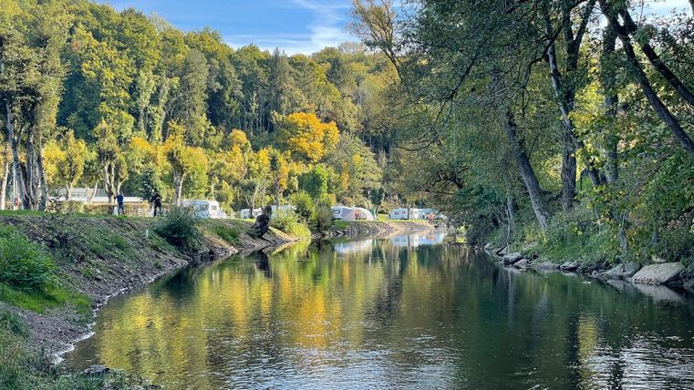 Een rustige rivier omgeven door kleurrijke bomen in de herfst. Op de achtergrond zijn enkele caravans langs de oever te zien.