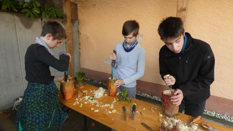 Trois garçons travaillent à une table pour couper et préparer des légumes. La table est couverte de différentes ingrédients et outils.
