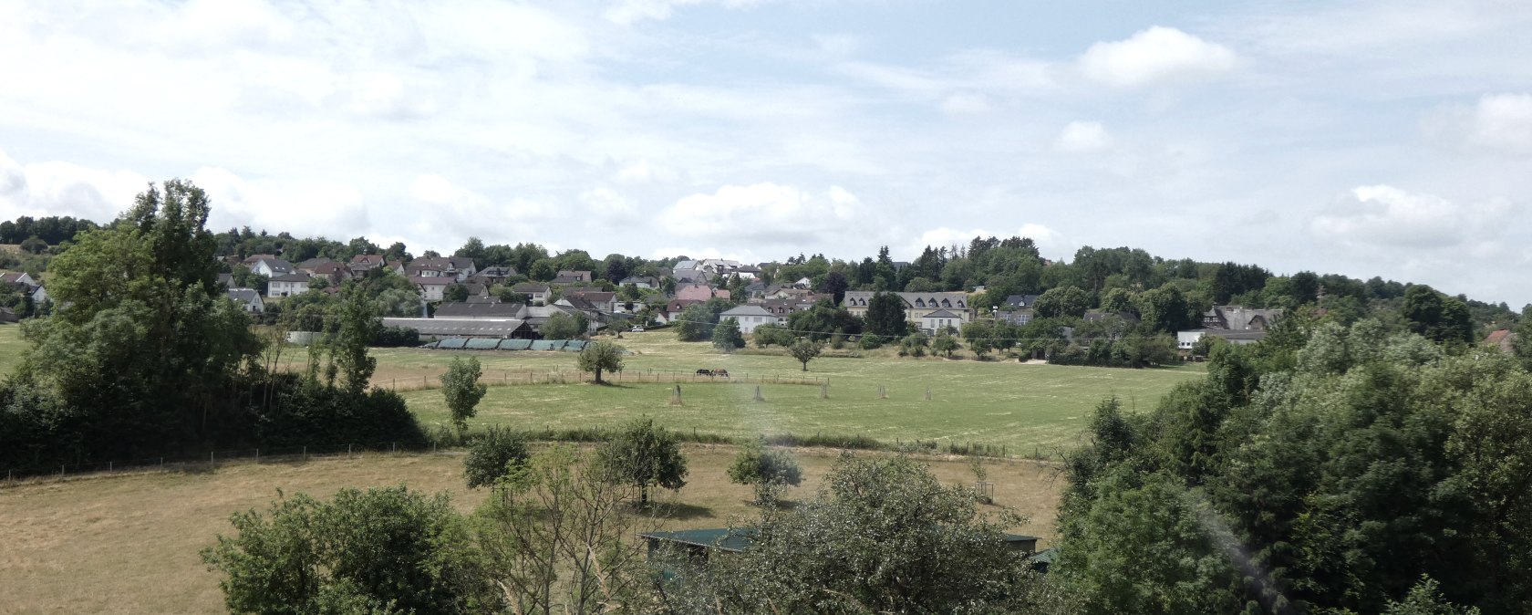 Grüne Wiesen und Bäume vor einem Dorf mit Häusern, unter einem blauen Himmel mit Wolken., © TI Bitburger Land