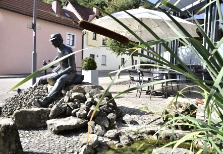 Bronze statue of a man on stones, surrounded by reeds and a small pond. Houses and a parasol can be seen in the background., © Tourist-Information Bitburger Land