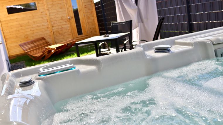 Un jacuzzi avec de l'eau bouillonnante est visible au premier plan. À l'arrière-plan, il y a un joli jardin avec une maison en bois et une table.