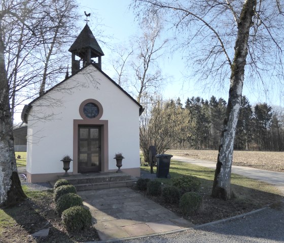 Kleine weiße Kapelle mit Glockenturm und Wetterhahn, umgeben von Bäumen und einem Feld. Der Himmel ist klar und blau., © Eifelverein OG Speicher