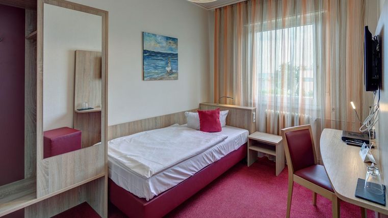 Ein modernes Hotelzimmer mit einem Einzelbett und einem kleinen Schreibtisch. Die große Fensterfront lässt viel Tageslicht herein und sorgt für eine angenehme Atmosphäre.