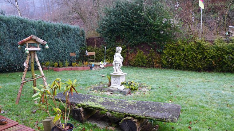 A quiet garden with a statue of a child in the middle. Surrounded by greenery and a wooden bench.