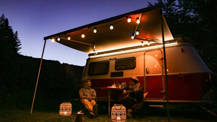 Ein gemütlicher Campingabend unter einem Zelt mit Lichtern. Zwei Personen sitzen entspannt neben einem Wohnwagen in der Natur.