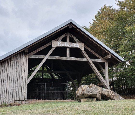 A wooden building with a 'lime kiln' sign stands in a green forest landscape. The sky is cloudy., © TI Bitburger Land S.Wagner