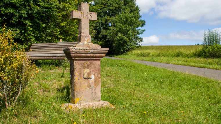 Ein steinernes Wegekreuz mit der Inschrift 'IHS' steht auf einer Wiese neben einem Weg, umgeben von Bäumen und einer Bank.