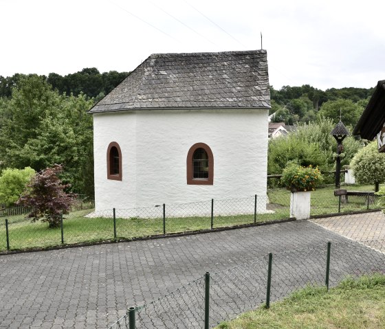 Weiße Kapelle mit Schieferdach, umgeben von grüner Landschaft. Ein Schild mit der Aufschrift "Kapellenhof" steht im Vordergrund., © TI Bitburger Land