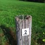 Un vieux poteau en bois avec un panneau indiquant le numéro 2 se trouve dans un pré vert. De l'herbe pousse sur le poteau., © Petra Fischer