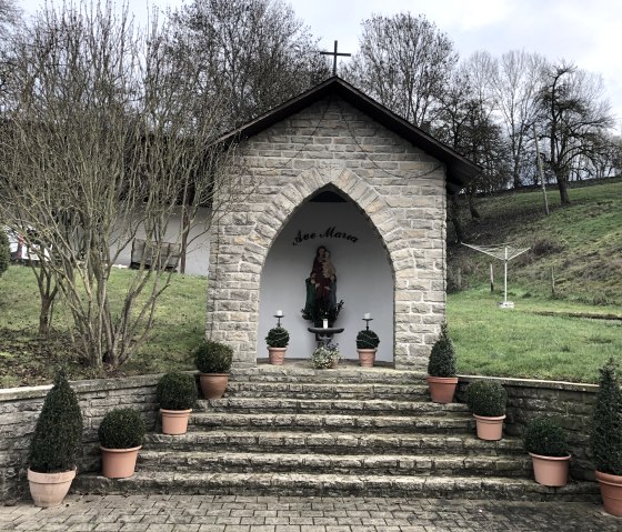 Kleine steinerne Kapelle mit Marienstatue, umgeben von Pflanzen in Töpfen. Die Kapelle steht auf einer Treppe, im Hintergrund Bäume und Wiese., © TI Bitburger Land - Biggi Thiebach