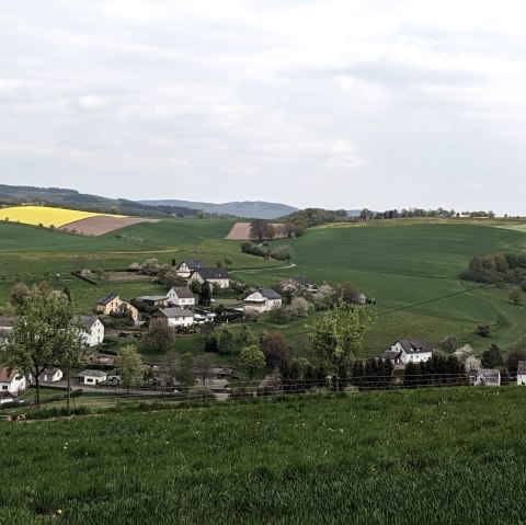 Groene heuvels met een geel koolzaadveld, een dorp in de vallei en een wandelaar op de voorgrond. Bewolkte lucht., © A. Girards