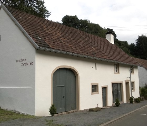 Weißes Gebäude mit der Aufschrift 'Kunsthaus Zendscheid', grünem Tor, umgeben von Bäumen und Straße. Personen sind im Vordergrund zu sehen., © R. Schaal
