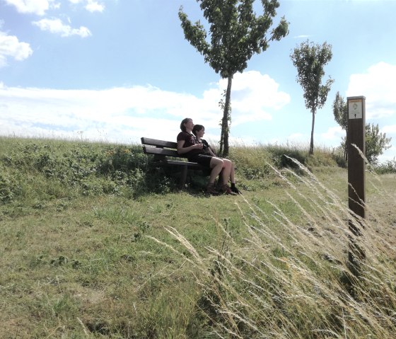 Two people are sitting on a bench in a green landscape. The sky is blue with a few clouds. A signpost is on the right of the picture., © Tourist-Information Bitburger Land