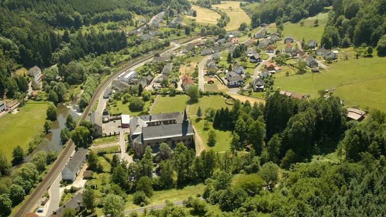 Luftaufnahme eines Dorfes mit Kirche, umgeben von Wäldern und Feldern.