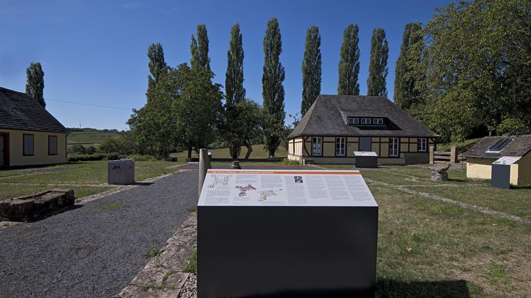 Eine historische Stätte mit mehreren Gebäuden und Stelen im Freien. Im Hintergrund stehen hohe Bäume unter einem klaren blauen Himmel.