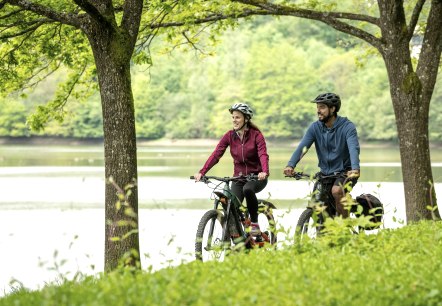 Zwei Radfahrer fahren lächelnd auf einem Weg am Stausee Bitburg entlang, umgeben von grüner Natur und Bäumen., © Eifel Tourismus GmbH, Dominik Ketz