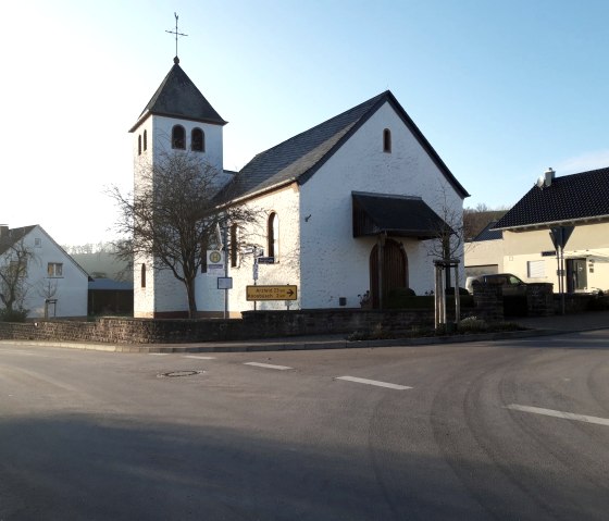 Eine kleine Kirche mit Turm in Wißmannsdorf, umgeben von Wohnhäusern, bei tiefstehender Sonne. Ein Wegweiser zeigt Richtung Arzfeld und Koosbüsch., © Georg Lotzkes