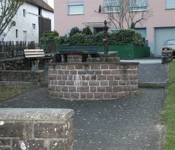 A village square in Gindorf with a stone fountain surrounded by benches. Houses and a parked car can be seen in the background., © Conny Meier