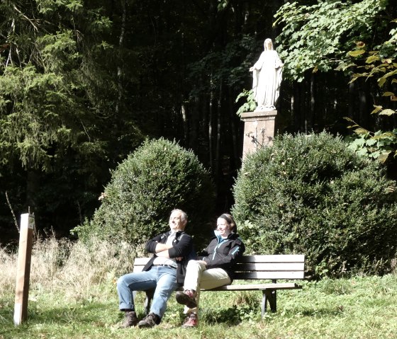 Twee mensen zitten op een bankje voor een Mariakolom in het bos. Ze genieten van de zon, omringd door struiken en bomen., © TI Bitburger Land