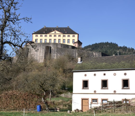 Kasteel Malberg troont op een heuvel, omringd door een stenen muur. Op de voorgrond staat een wit huis met een groen dak., © TI Bitburger Land
